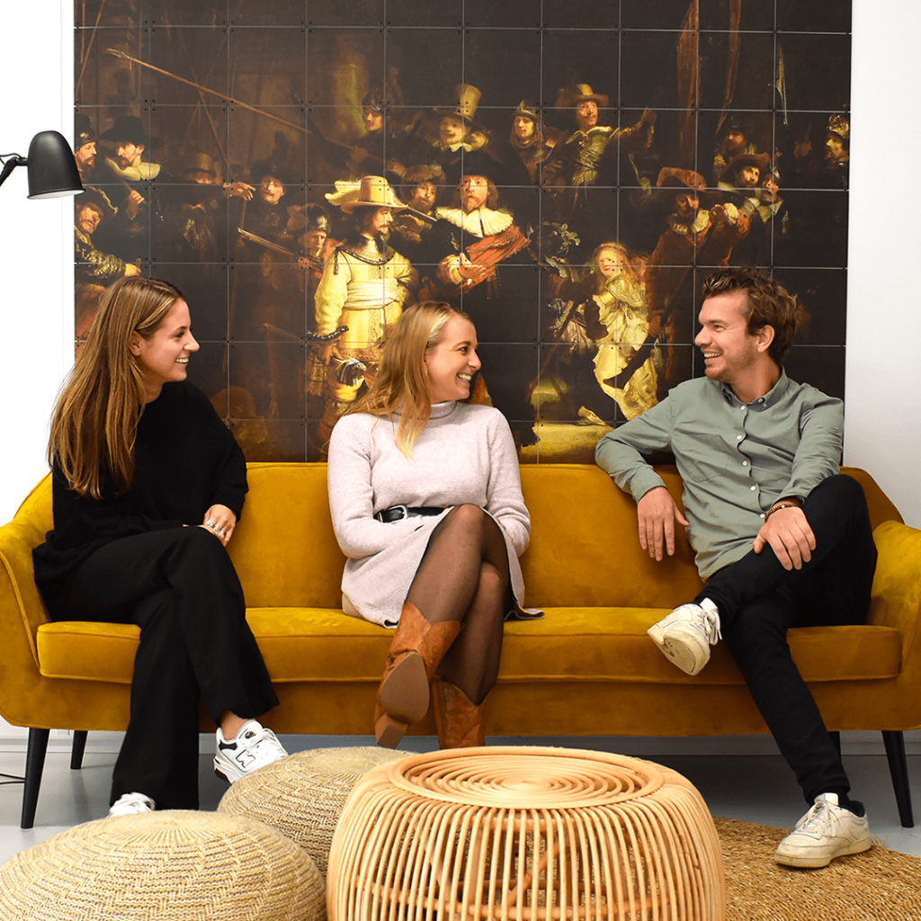 Twee vrouwen en een man op een gele bank met de Nachtwacht achter zich